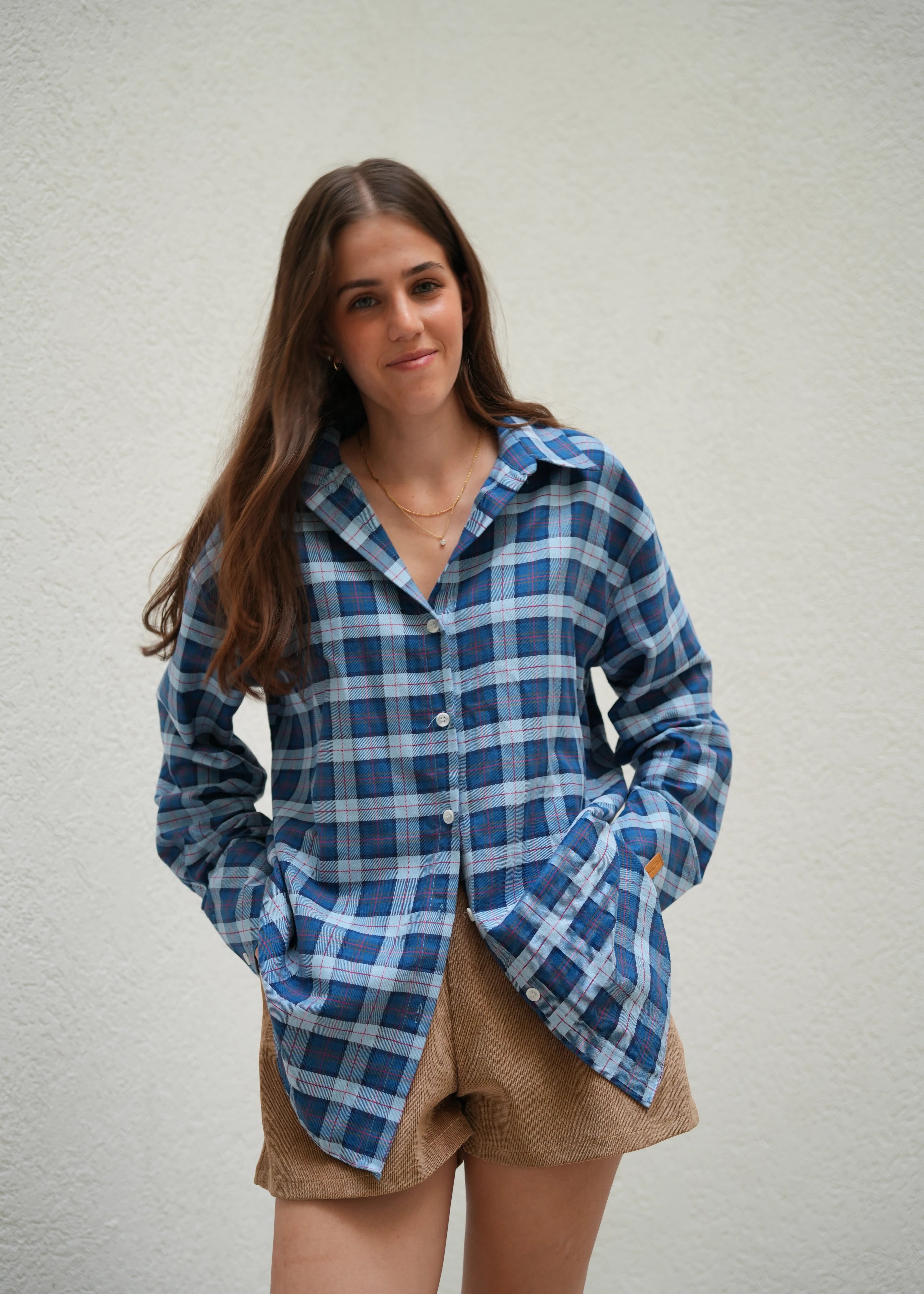 Person wearing a blue plaid shirt with a brown jacket over shoulders on a light gray background